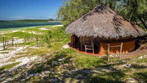 A thatched-roof hut with a wooden door and a small porch, surrounded by a sandy beach and a clear view of the ocean. - Varatraza Wind Riders (Antsiranana)