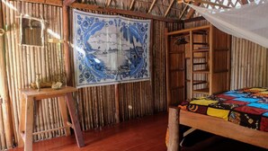 A room with a wooden bed, a small table with a vase, a hanging tapestry, and a bamboo wall. - Varatraza Wind Riders (Antsiranana)