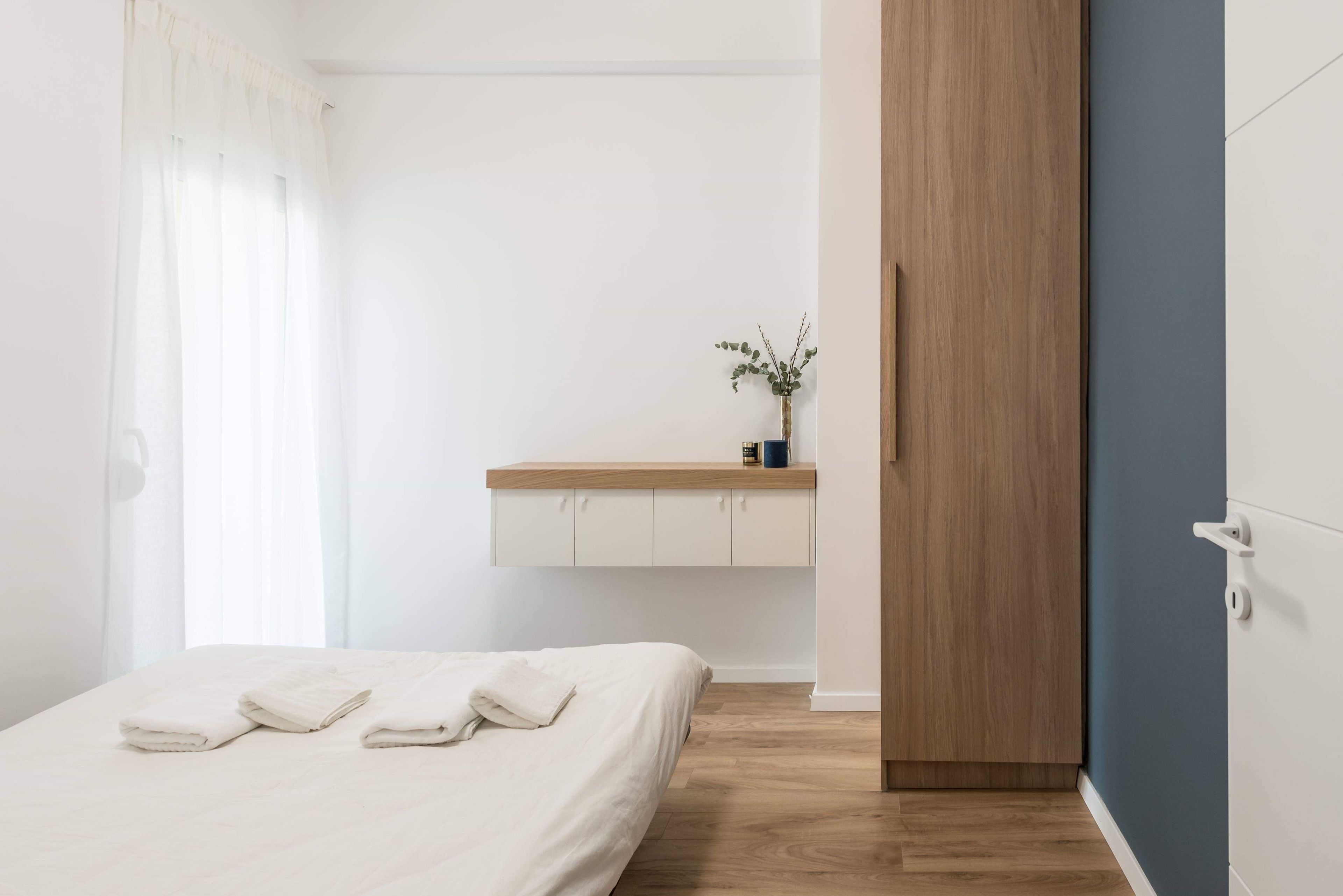 A bedroom with a bed, a wooden wardrobe, a small shelf with a vase, and a window with curtains.
