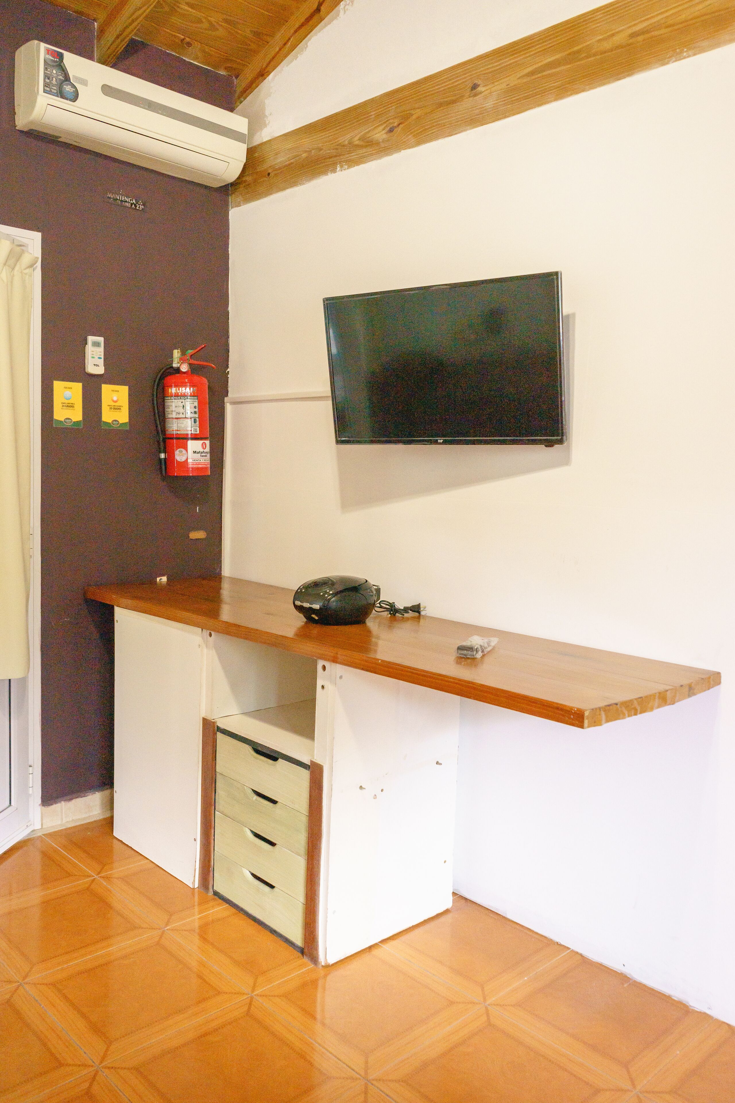 A room with a wooden desk, a mounted television, a fire extinguisher, and a wall-mounted air conditioning unit.