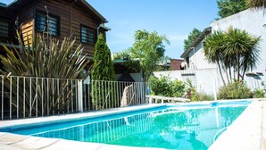 Una piscina con cerramiento metálico, rodeada por una casa de madera y un edificio blanco.