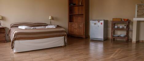 A bedroom with a bed, a wooden wardrobe, a small refrigerator, and a shelf with dishes.
