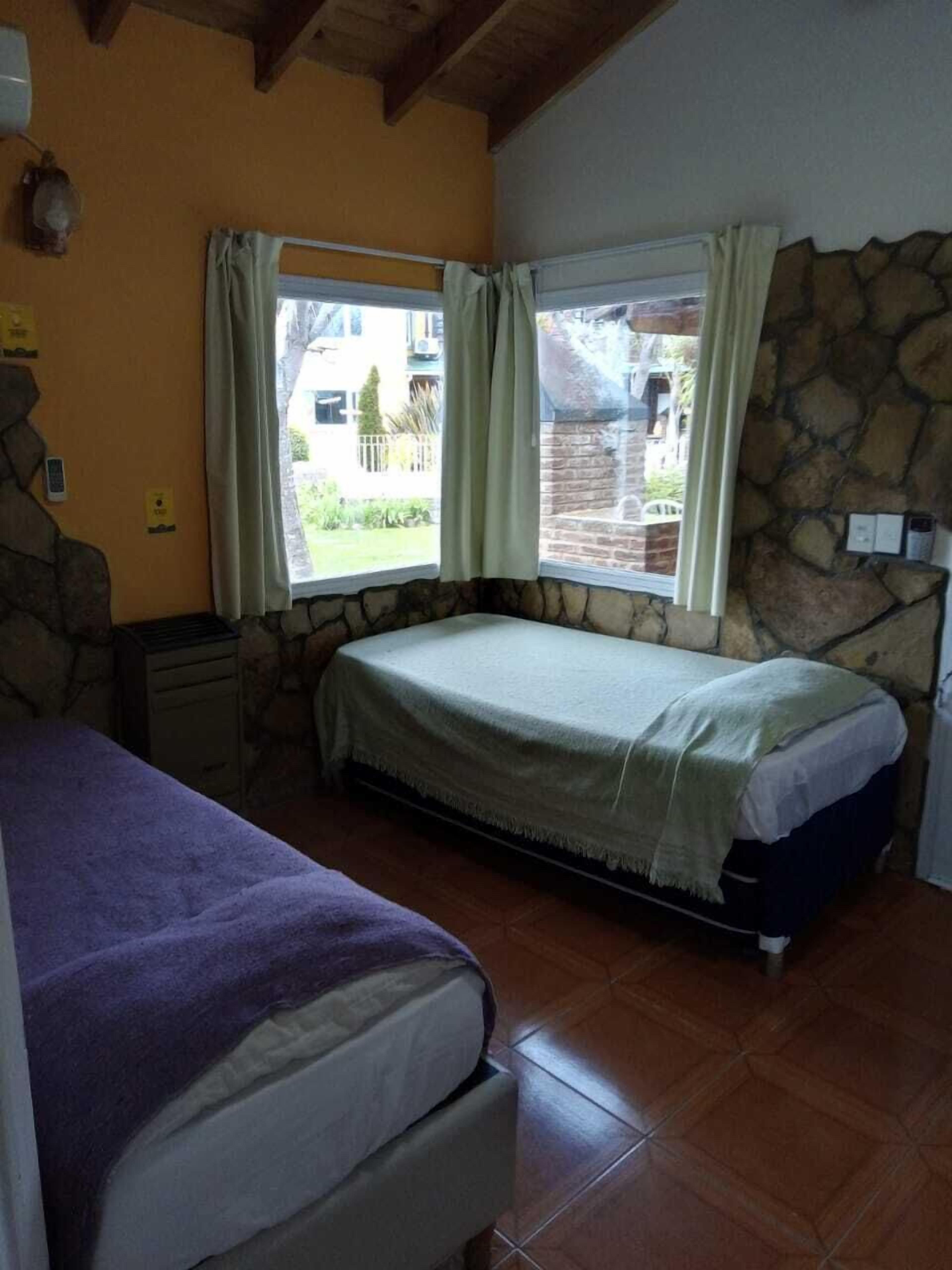 A bedroom with a bed, a nightstand, a window with curtains, and a stone wall.