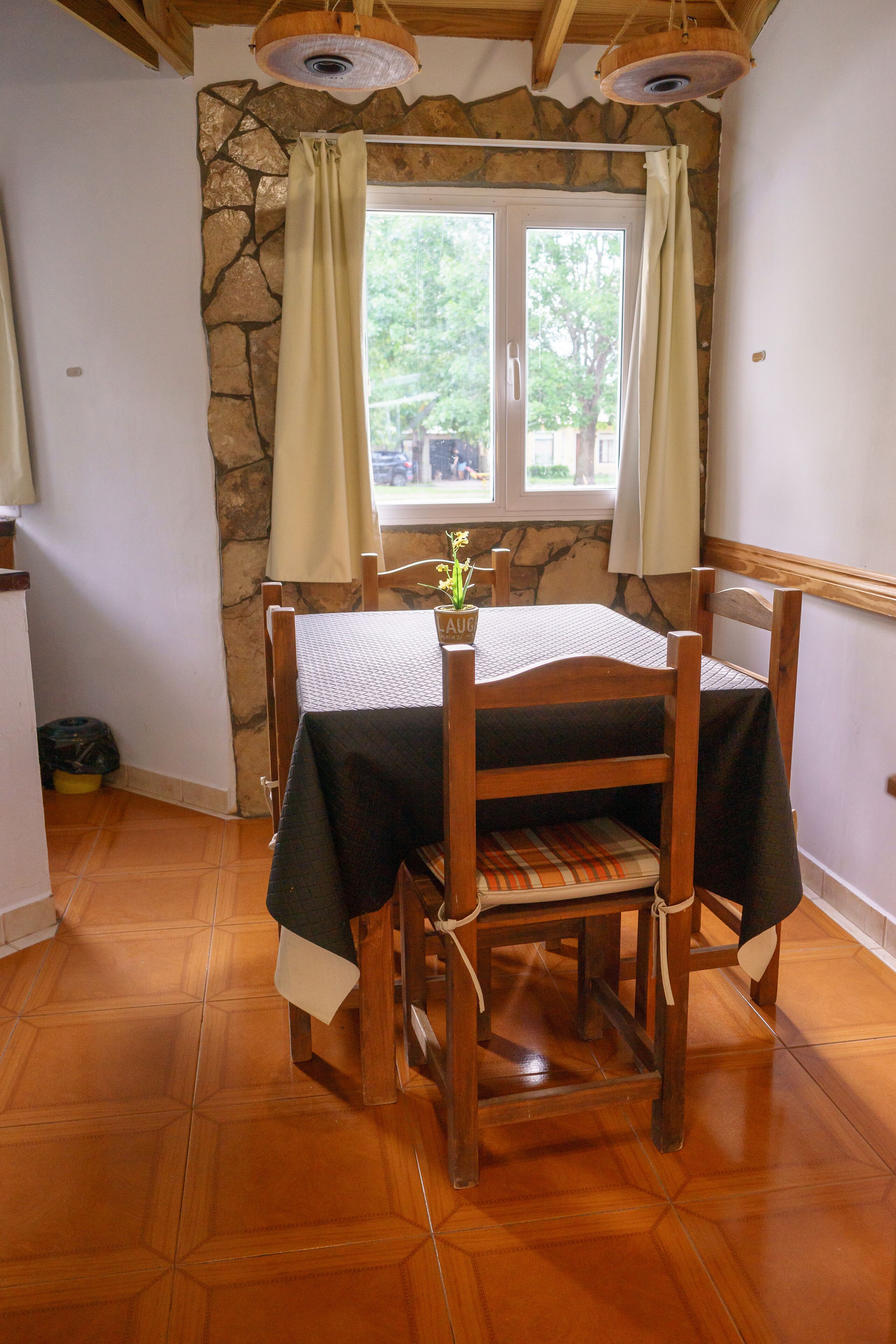 Un comedor con pared de piedra, mesa de madera, sillas y ventana con cortinas.