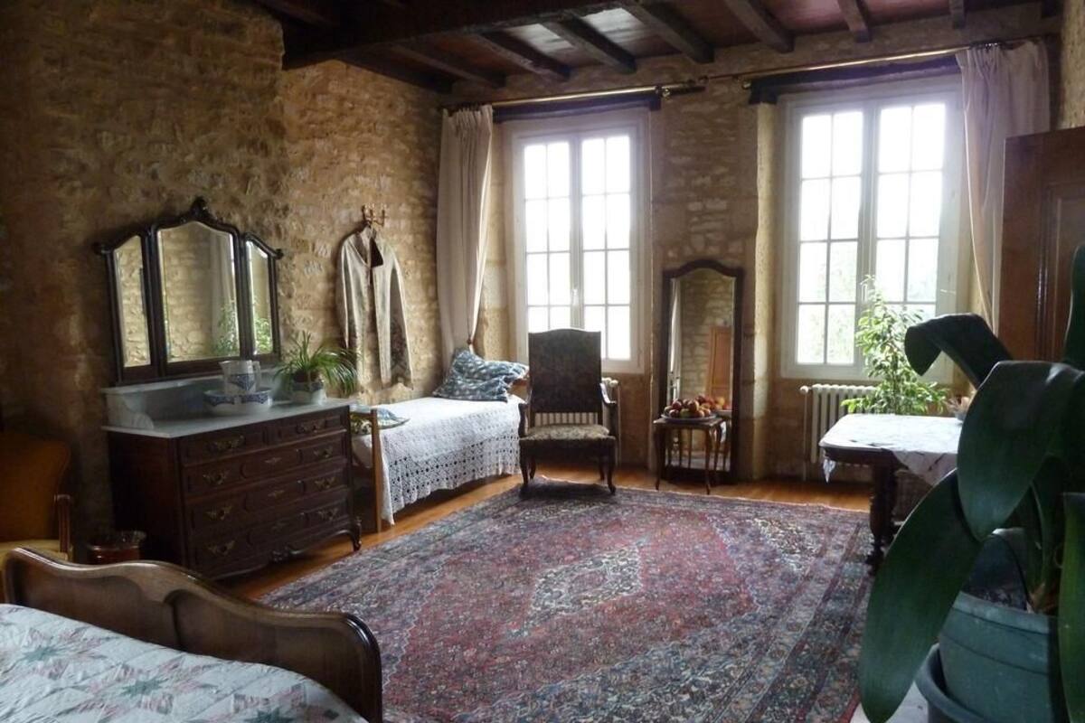 Un dormitorio rústico con una cama grande, un aparador, una silla y una mesa. Hay dos ventanas con cortinas, un espejo y una planta en maceta.