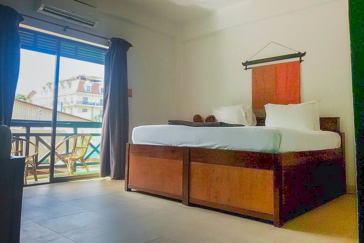A hotel room with a bed, a small table, and a balcony with chairs.