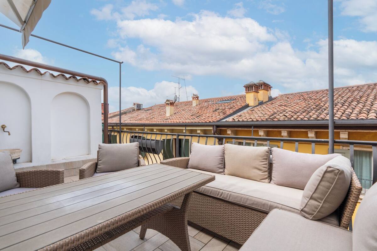 A patio with wicker furniture and a table, overlooking a cityscape with terracotta roofs.