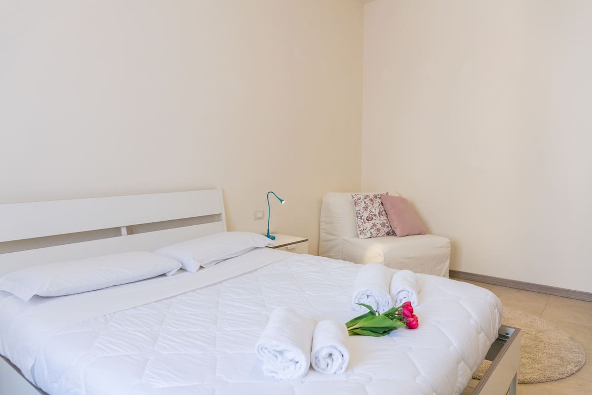 A neatly made bed with white linens and rolled towels, a bedside table with a lamp, a chair with a floral pillow, and a small round rug on the floor.