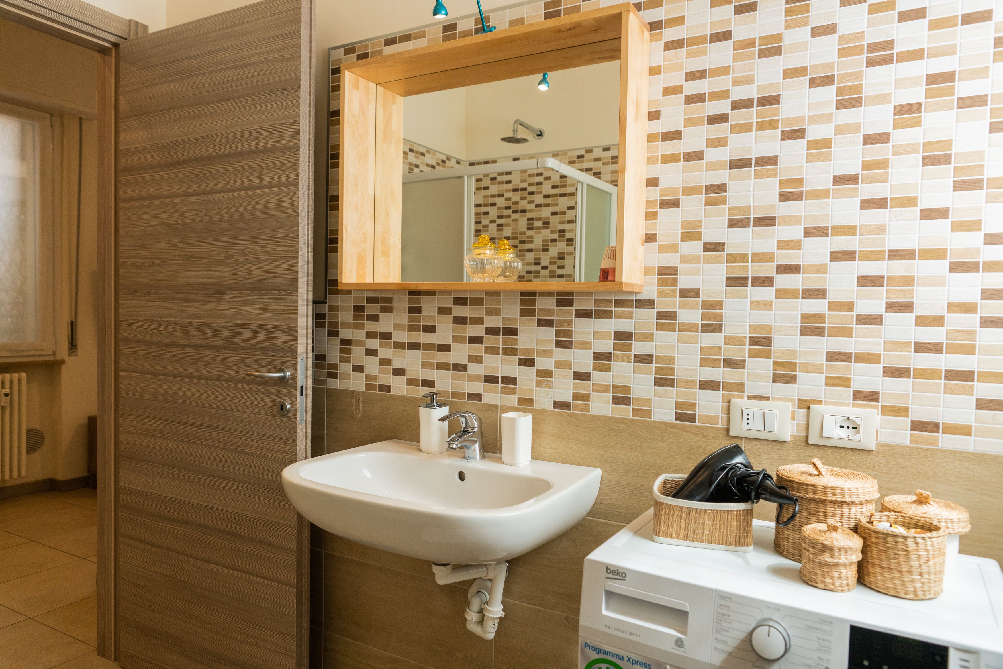 Un baño con puerta de madera, un lavamanos con espejo arriba, pared de azulejos y un electrodoméstico blanco en la encimera.