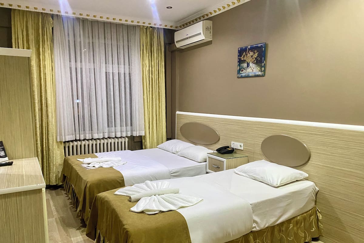 A hotel room with two beds, a bedside table, an air conditioning unit, and a painting on the wall.