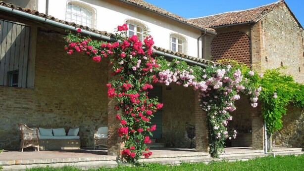 A stone building with a tiled roof, a covered porch with pink flowers, and a bench.