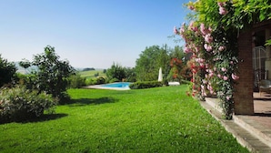A well-maintained garden with a swimming pool, lush greenery, and a brick building with a pergola covered in flowers.
