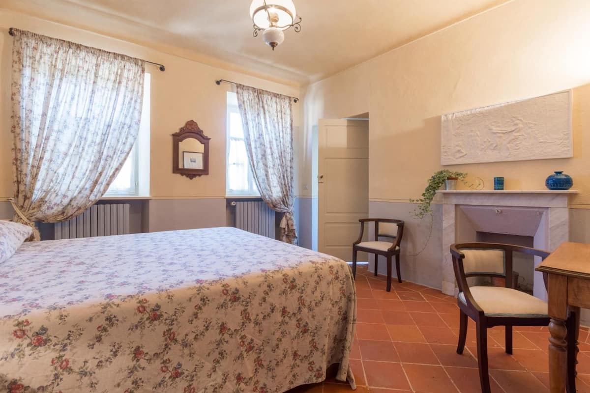 A bedroom with a bed, two chairs, a fireplace, and a window with curtains.