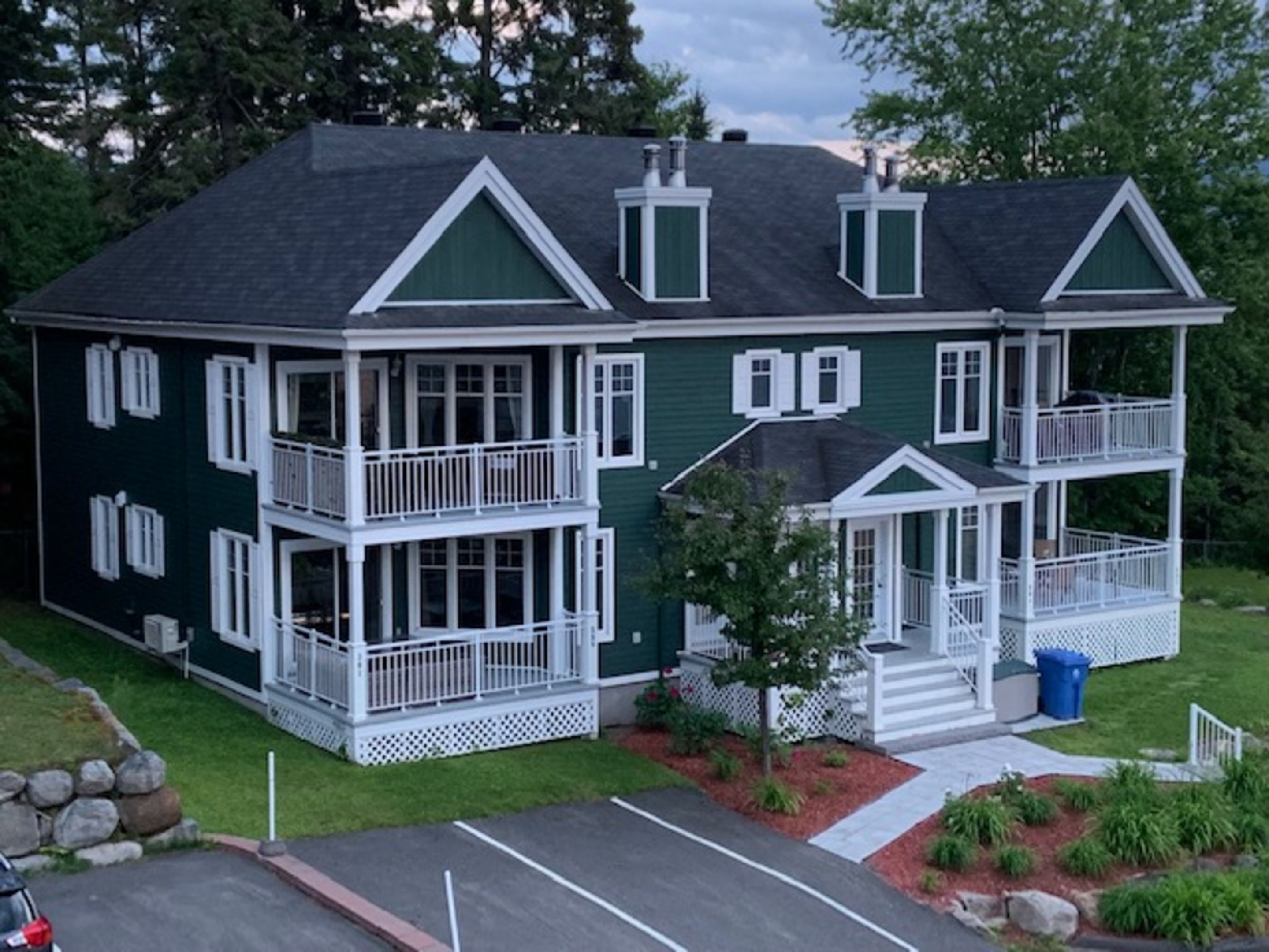 A two-story house with a balcony on the second floor, a front porch, and a small front yard with a tree and a trash bin.