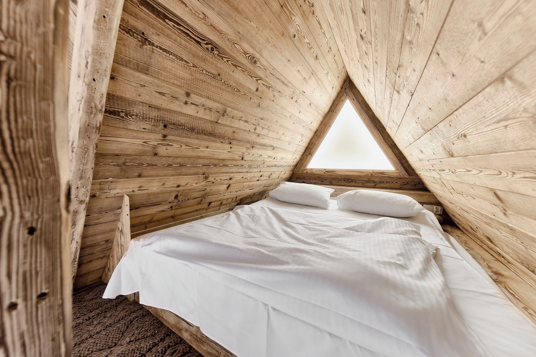 A wooden interior with a triangular window, a bed with white bedding, and a wooden floor.