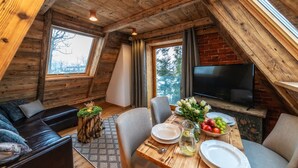 A cozy room with a wooden ceiling, a TV mounted on the wall, a dining table set with plates, glasses, and a vase of flowers. - Hel Apartments (Zakopane)