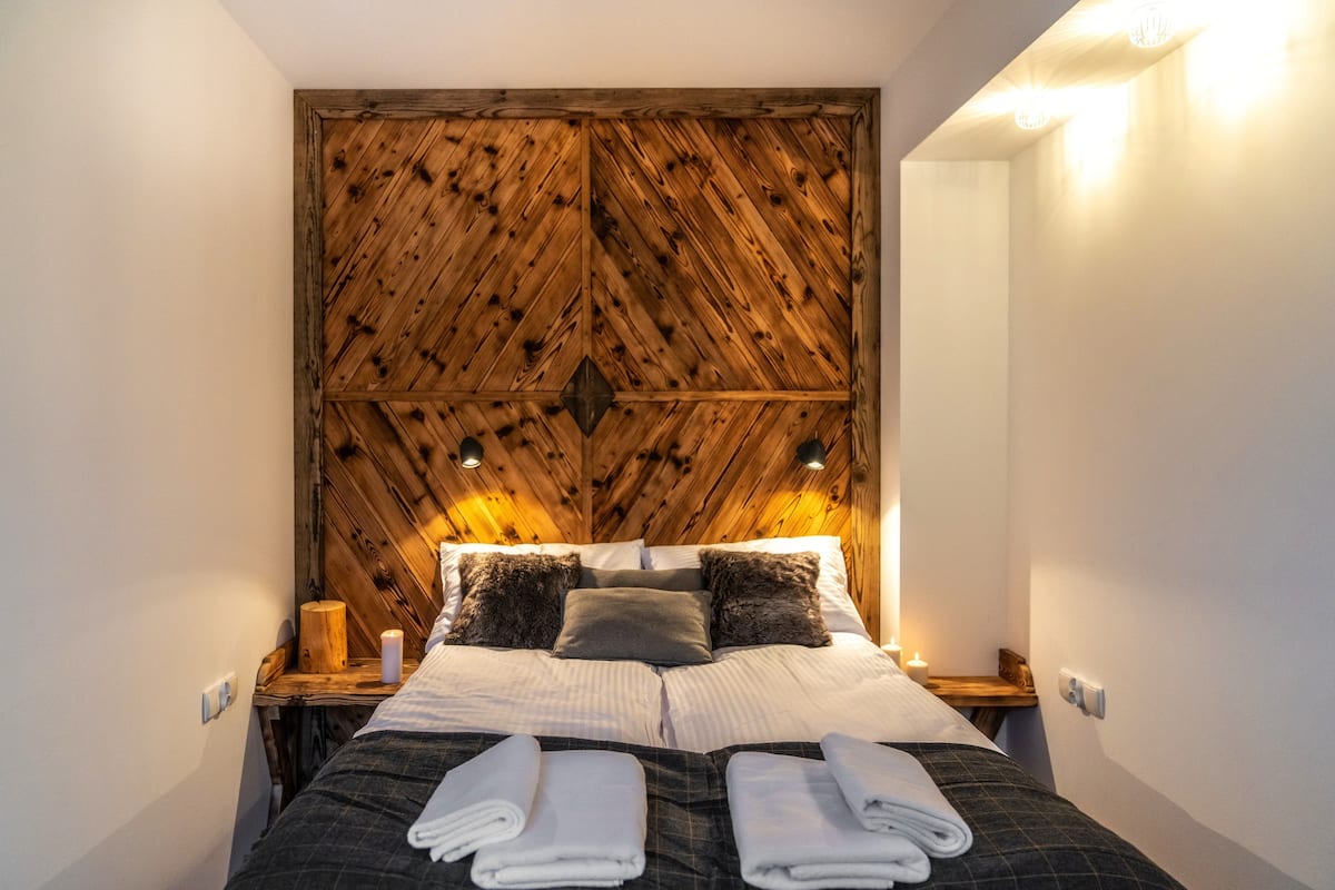 Una cama bien hecha con cabecera de madera, dos mesitas de noche y una pequeña mesita con una vela.