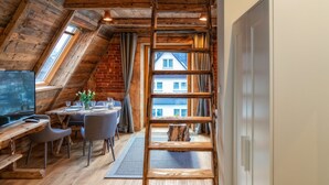A rustic room with a sloped ceiling, wooden beams, a dining area with a table and chairs, and a large window overlooking a building. - Hel Apartments (Zakopane)