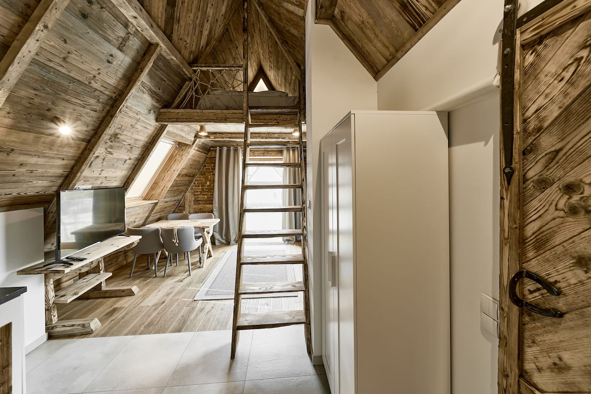 Un cuarto estilo loft con techo de madera, una escalera que lleva a un nivel superior, un comedor con mesa y sillas, y un refrigerador blanco.