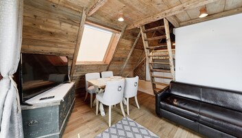Et loftrum med træloft, spisebord med stole, en sort lædersofa og en stige til en alkove.