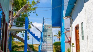 Un étroit sentier mène à un escalier doté d’une balustrade en verre, bordé de bâtiments aux façades bleues et blanches, et s’ouvre sur un petit espace jardin avec un arbre et des fleurs.