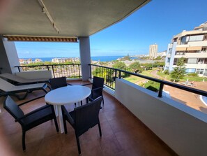 A balcony with a table and chairs, overlooking a coastal view with buildings and greenery.