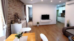 A modern living room with a TV mounted on the wall, a dining area with a wooden table and white chairs, and a kitchen in the background. - Rialto Luxury Apartment (Sibenik)
