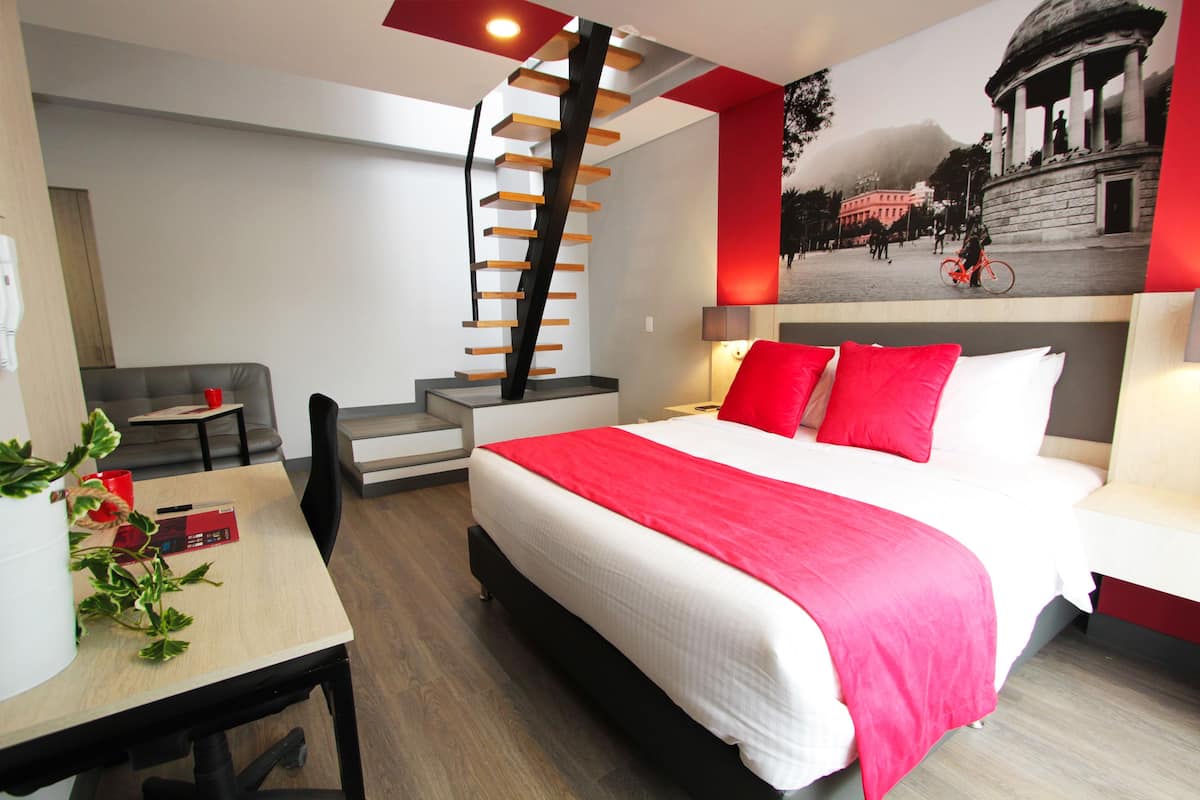 A modern hotel room with a large bed, a staircase, a desk, and a dining area.