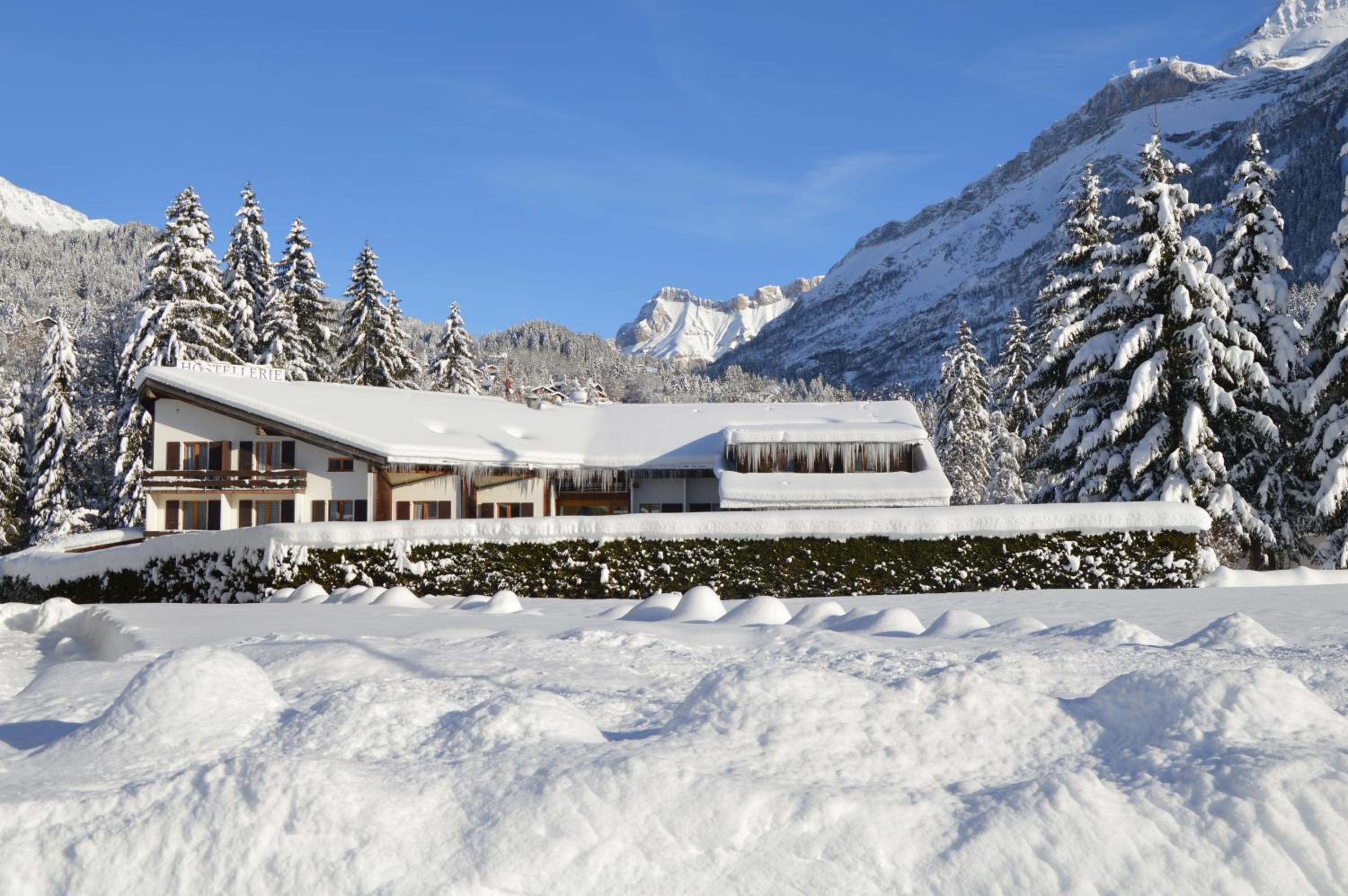 Un lodge de montaña cubierto de nieve con un techo grande y múltiples ventanas, rodeado de árboles y montañas cargadas de nieve.