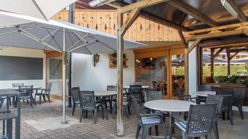 Un patio al aire libre con mesas y sillas, vigas de madera y un área cubierta con sombrillas.