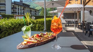 A table with a glass of orange cocktail, a bottle of water, and a wooden board with various food items. - Hôtel Les Sources (Ormont-Dessus)