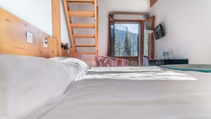 A bedroom with a wooden headboard, a bed with white linens, a striped chair, a desk with a lamp, and a TV on the wall. - Hôtel Les Sources (Ormont-Dessus)