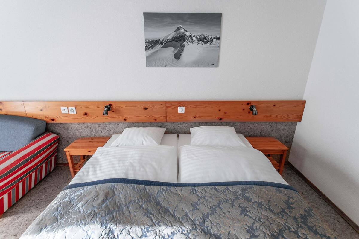 A bedroom with a bed, two bedside tables, a wooden headboard, a red striped bench, and a mountain landscape painting on the wall.