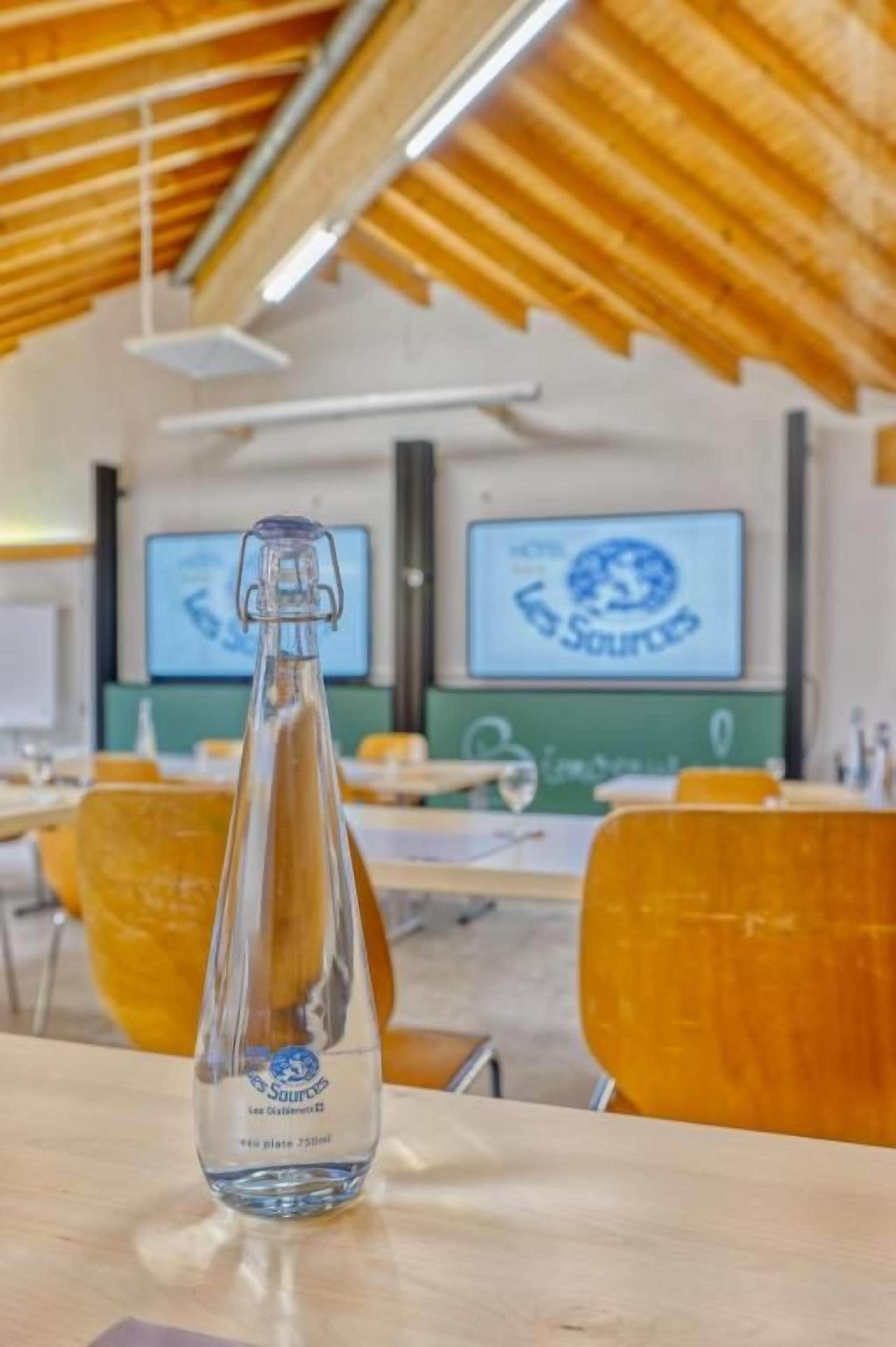 A bottle of water on a table in a classroom with wooden desks and chairs, and two screens displaying the word 'SOURCES'.