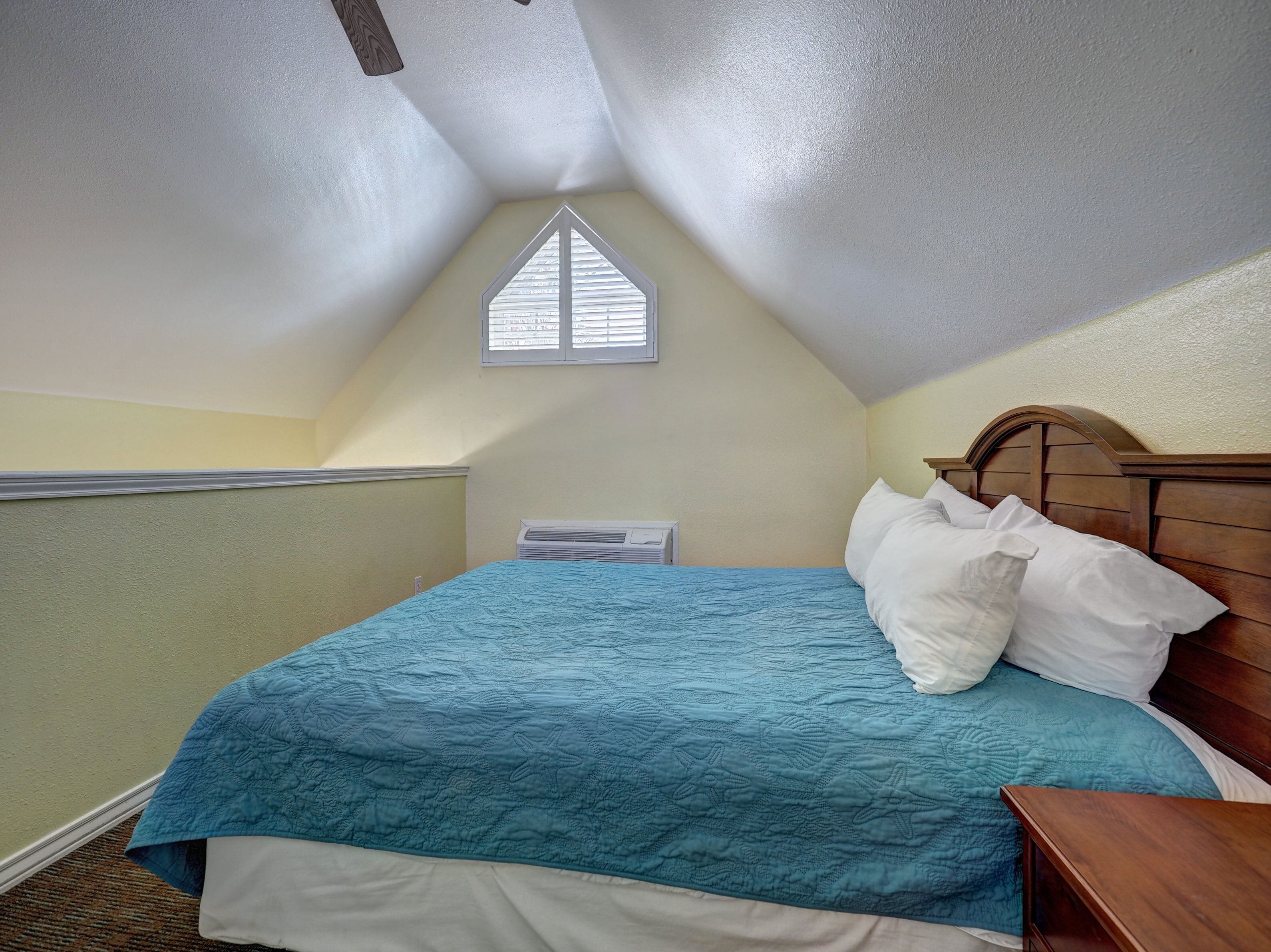 Een slaapkamer met een schuine plafond, een raam, een bed met een blauwe quilt en een houten hoofdbord.