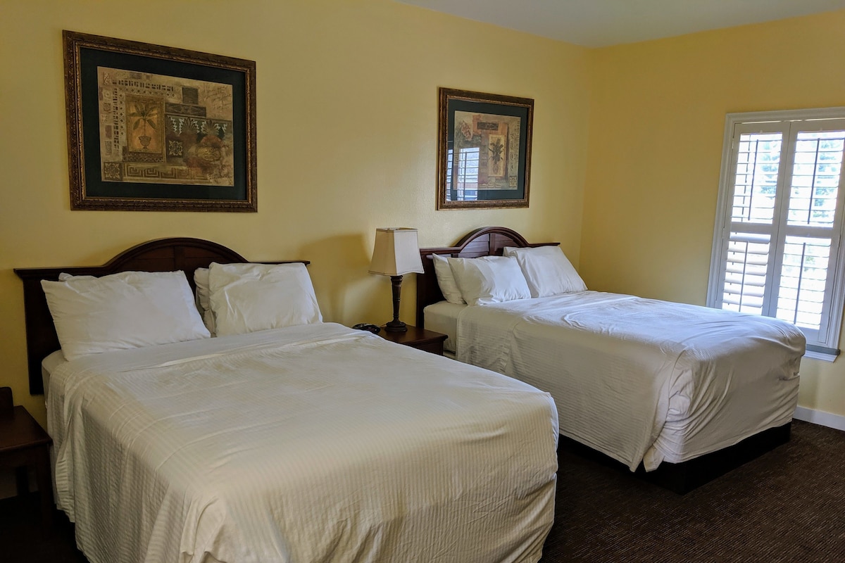 A hotel room with two beds, a nightstand, a lamp, and two framed pictures on the wall.