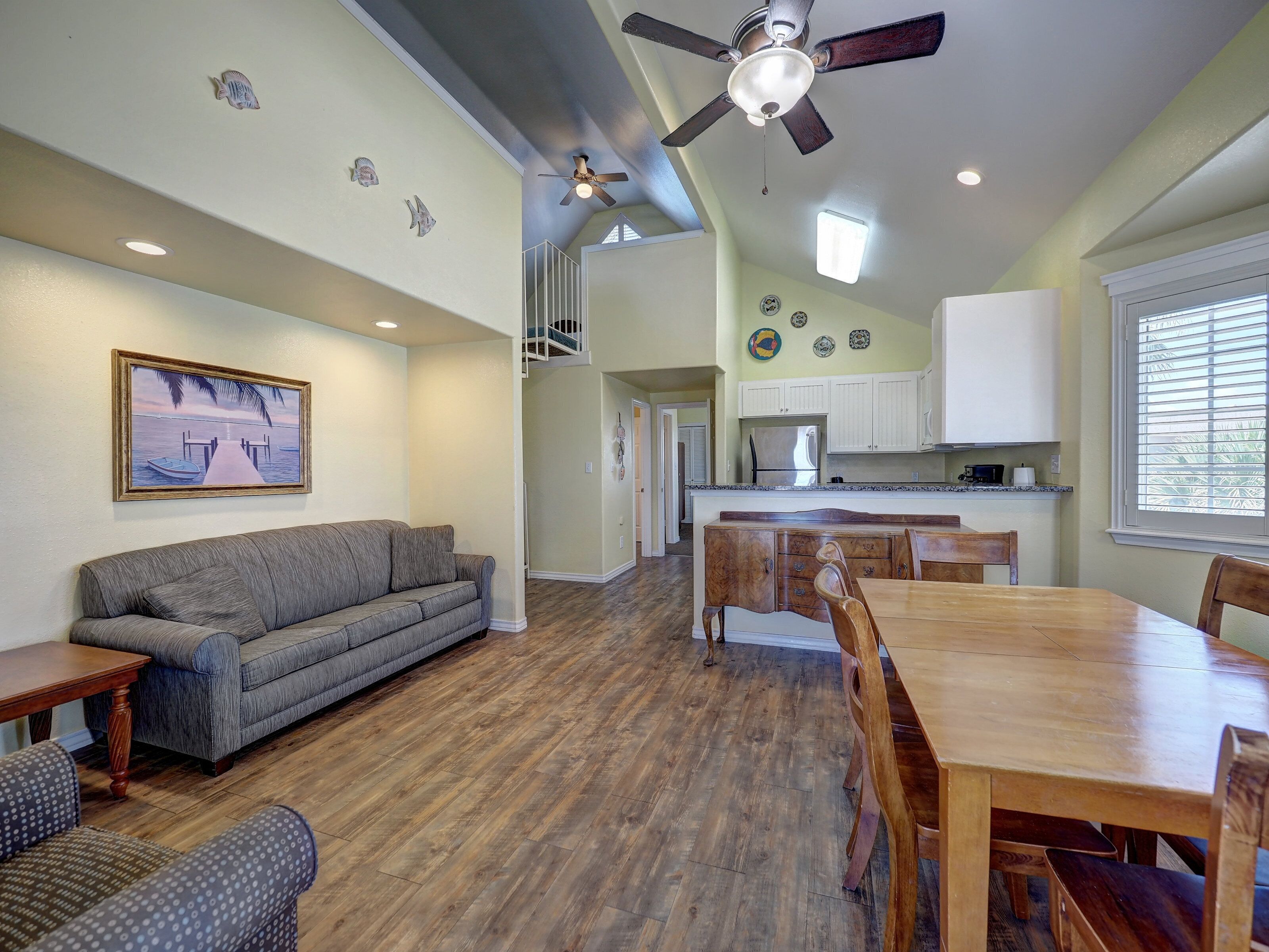 A spacious living area with a gray sofa, wooden dining table, and chairs. A ceiling fan and recessed lighting are visible.