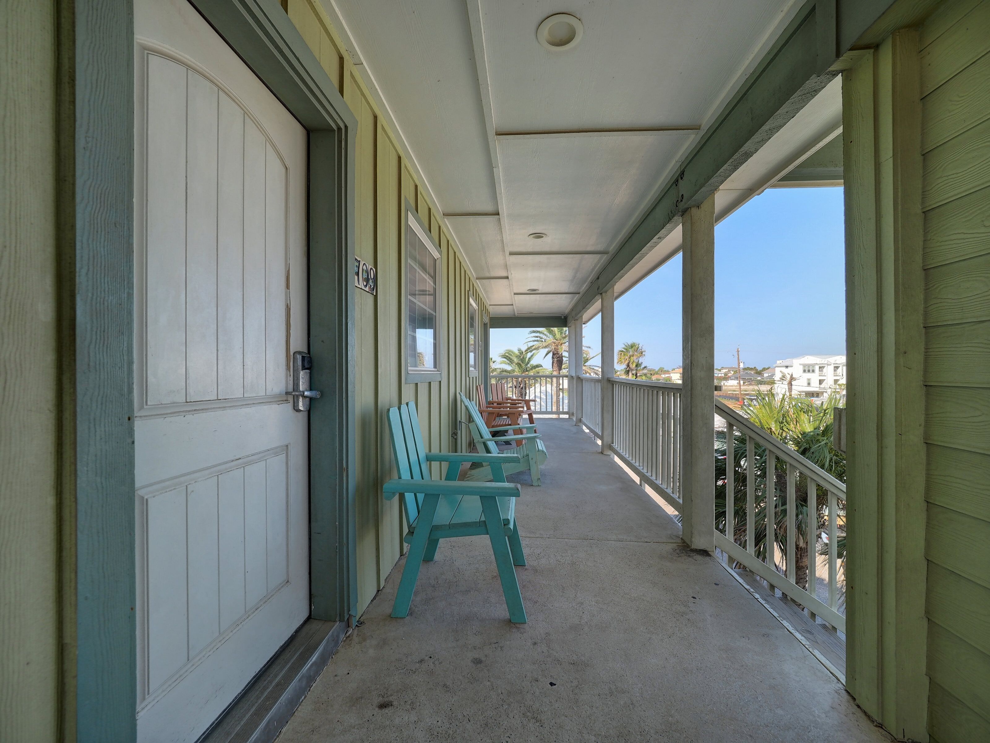 A covered balcony with teal chairs, a door with a number sign, and a view of palm trees and buildings.