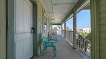 A covered balcony with teal chairs, a door with a number sign, and a view of palm trees and buildings.