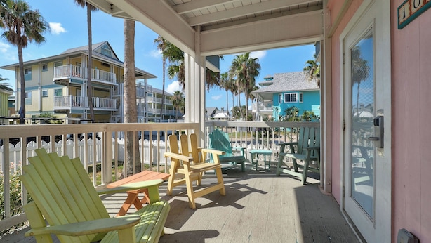 A covered outdoor area with wooden chairs and a table, a white picket fence, and a view of nearby buildings. - Seashell Village Resort near the beach with kitchens (Port Aransas)