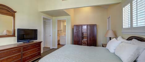Un dormitorio con cama, cómoda, televisor, armario y un baño visible a través de una puerta abierta.