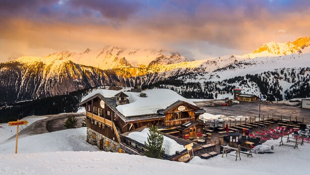 Eine Holzhütte mit verschneitem Dach, einem Essbereich mit Außenplätzen und Blick auf verschneite Berge unter einem Abendhimmel.
