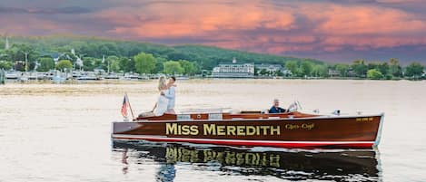 A boat named 'Miss Meredith' with a flag, docked near a waterfront with buildings and trees in the background.