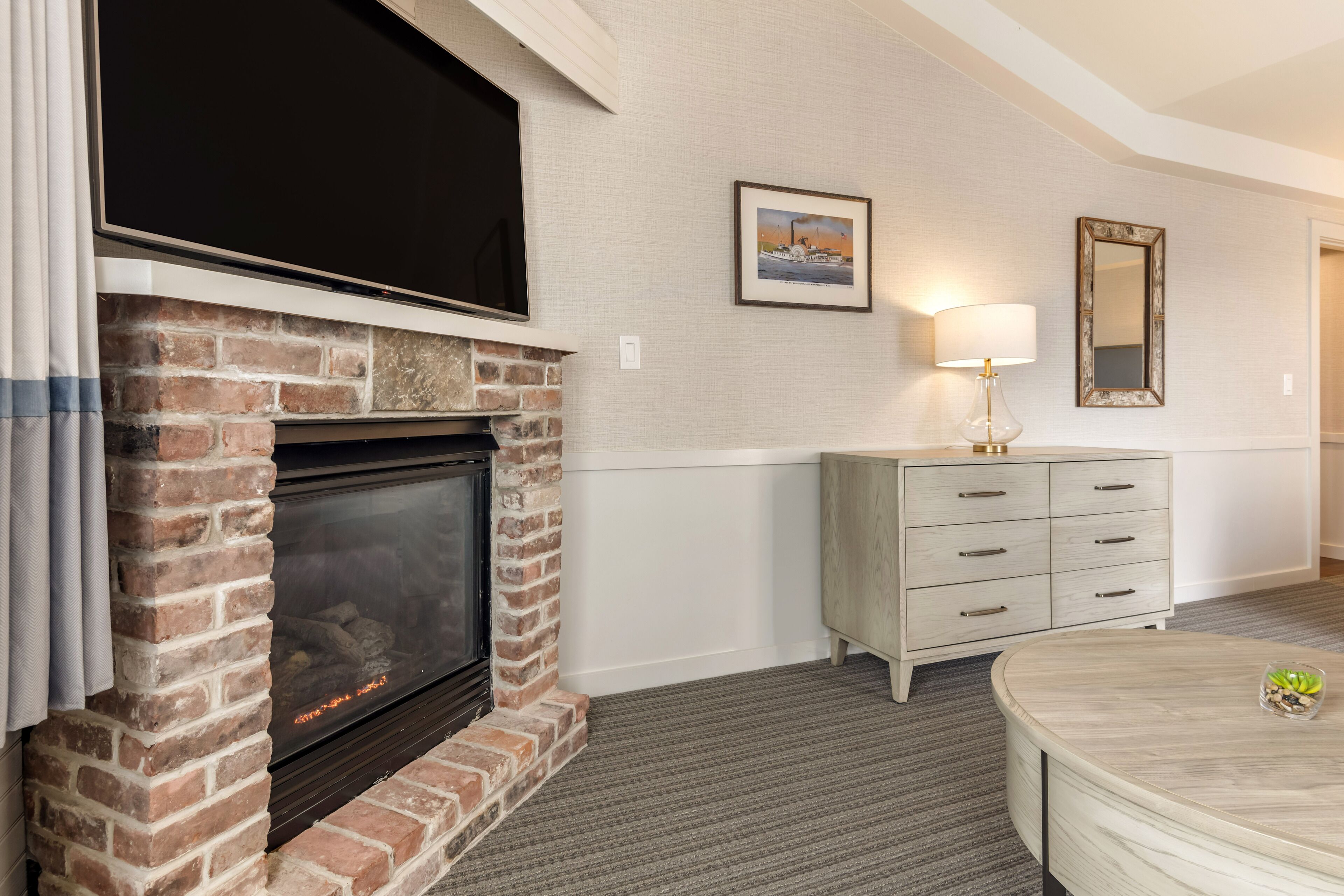 A room with a brick fireplace, a flat-screen TV mounted above it, a light-colored dresser, and a round table with a vase.
