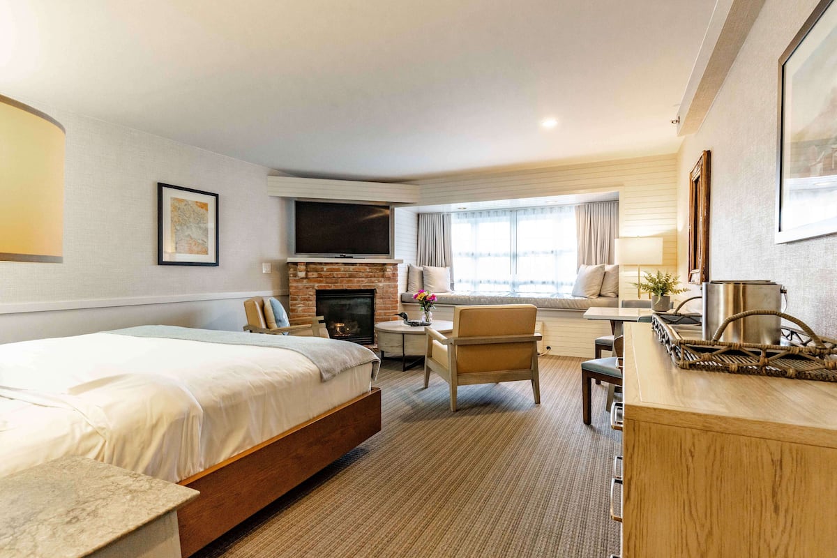 A hotel room with a large bed, a desk, a chair, a TV, a fireplace, and a window with curtains.