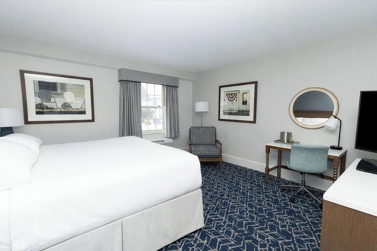A hotel room with a large bed, a desk with a chair, a round mirror, and two framed pictures on the wall.