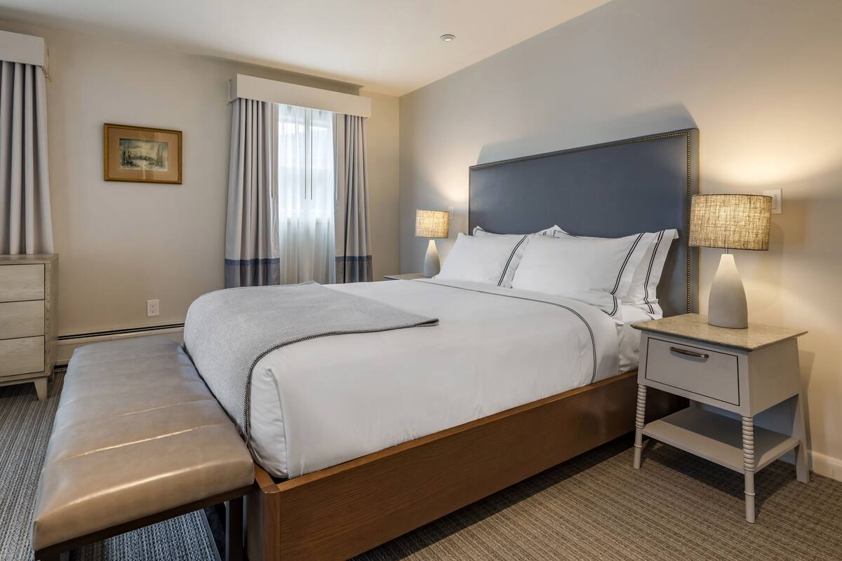 A hotel room with a large bed, a bench, two bedside tables with lamps, a wall-mounted picture, and a window with curtains.