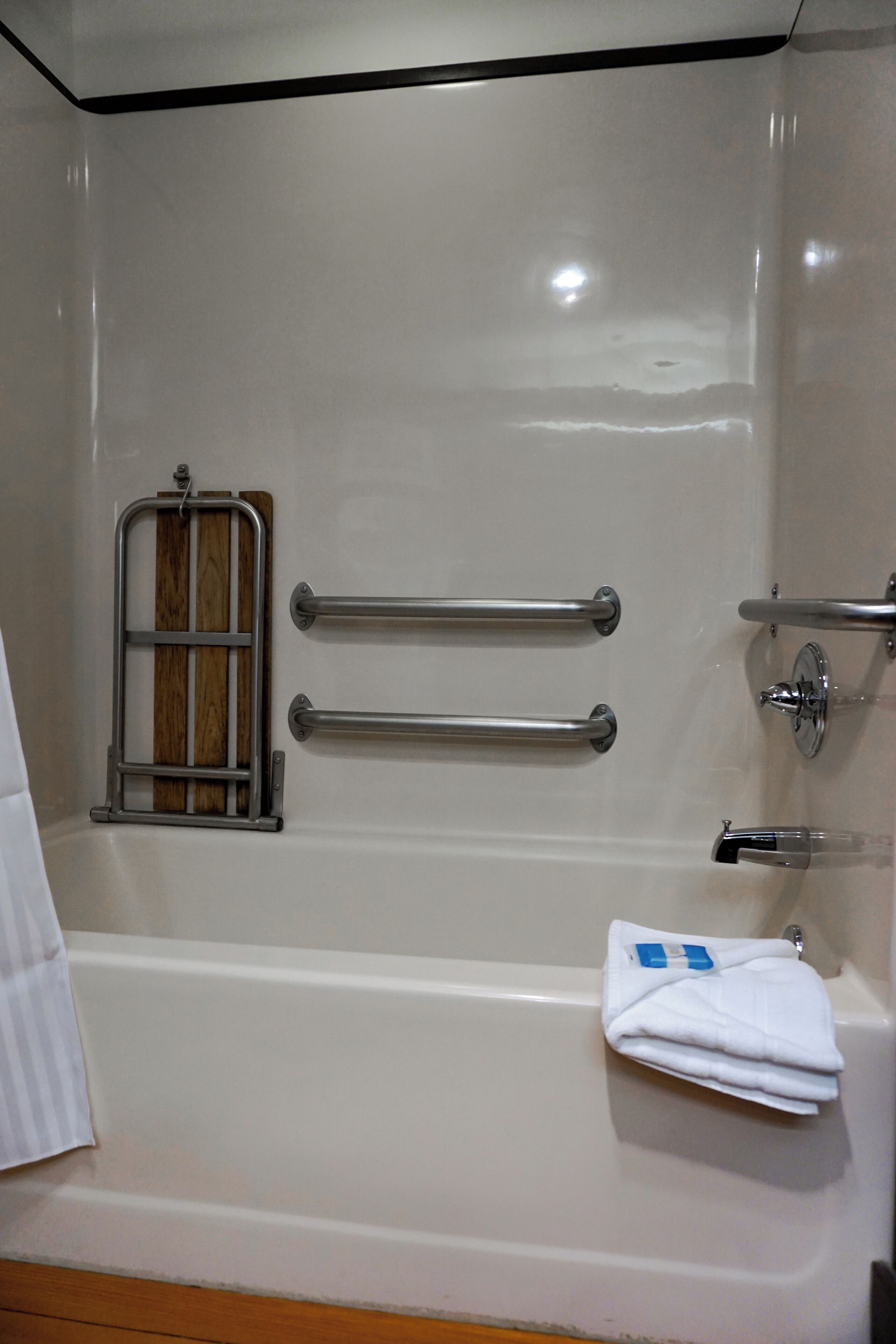 Une salle de bain avec une baignoire blanche, deux barres d’appui, un panneau en bois et une serviette blanche pliée.