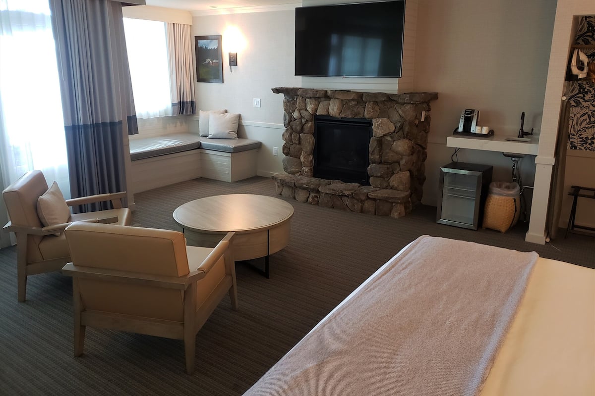 A hotel room with a stone fireplace, a flat-screen TV, a seating area with a bench, and a round table.