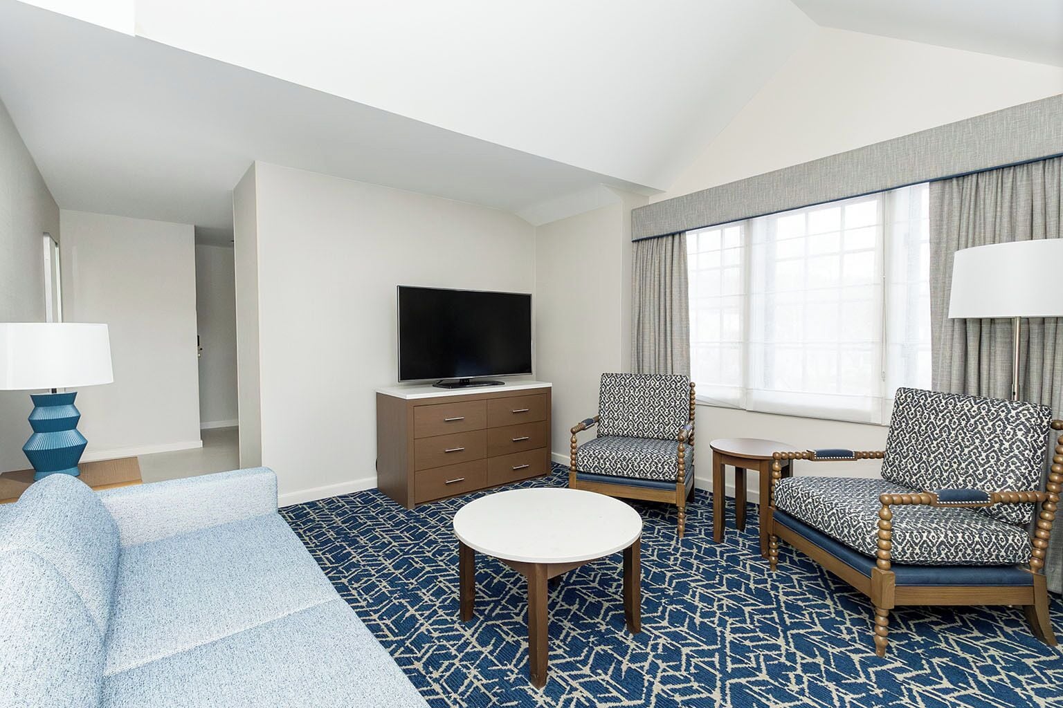 A hotel room with a sofa, two patterned armchairs, a small table, a TV on a wooden cabinet, and a window with curtains.